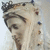 A pixelated figure with long light hair and a dark crown-like headpiece, wearing a white veil or shawl against a pale background.