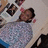 A person in a gray sweater and blue pants sits at a table with multiple framed photos, holding a pink object near their face.
