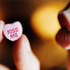 A hand holds a small pink heart-shaped candy with red text reading 'KISS ME' against a blurred warm-toned background.