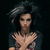 A person with voluminous dark hair stands against a black background, hands crossed in front of chest, wearing a dark shirt with floral pattern.