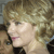 Pixelated close-up of a person with light brown curly hair, wearing a dark top, against a blurred background.