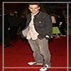 A person stands on a red carpet wearing a dark denim jacket, light patterned shirt, gray pants, and white sneakers with black accents.