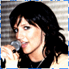 A woman with dark shoulder-length hair holds a silver microphone, wearing a black top and silver necklace against a striped background.
