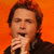 Pixelated image of a person with brown hair holding a microphone against an orange background.
