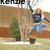 A person in a blue top and light blue jeans jumps on a brown bench outdoors with trees and a building background, text 'Kenzie' appears below.