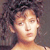 Pixelated static image of a person with curly dark hair and light skin tone, cropped to show upper shoulders and head against a gray background.