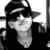 Black-and-white image of a person wearing a cap and sunglasses, with a neutral expression, displayed in a