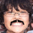 A person with dark curly hair and glasses smiles broadly, showing teeth and a mustache in three