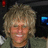 Pixelated static image of a person with light brown curly hair, closed eyes, and a neutral expression against dark background elements.