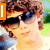 A pixelated image of a person with brown curly hair and sunglasses, wearing a white top against a blurred green background.