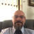 A pixelated static image shows a person with a bald head, dark beard, and mustache wearing a light-colored shirt and dark tie, seated against a blurred background with vertical lines and a framed item on the wall.