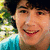 A person with dark curly hair wearing a blue shirt smiles in a pixelated outdoor setting with green foliage background.