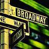 A street signpost with multiple directional signs showing 'BROADWAY', 'ONE WAY', and partial text on a yellow pole against blurred green background.
