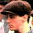 Pixelated close-up of a person wearing a dark flat cap, light-colored shirt, and dark tie, with visible skin tones and blurred background elements.