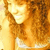 A woman with long curly brown hair wears a white top, smiling in a warm-toned image with visible pixelation.