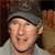 Pixelated close-up of a person wearing a gray cap and light-colored shirt, with a blurred background showing brown and dark tones.