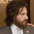Pixelated image of a person with long brown hair and a beard, wearing a white shirt under a dark jacket, positioned against a brown background.