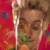 Pixelated static image of a person with light brown hair holding a microphone against a red background, featuring green and orange color accents on the lower portion.