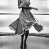 Black-and-white image of a woman in a flowing dress with a black belt, holding a bag, standing against a blurred background.