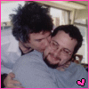 Two people embrace in a room with light-colored walls and windows; one wears a white shirt, the other a gray shirt. A pink border frames the image.