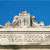 Pixelated image of a white architectural structure with columns and a central dome against a solid blue background, with a horizontal band of indistinct text below.