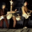 Animated sequence of two musicians playing electric guitars on a dark stage with a wooden floor, captured in a of