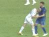 Two soccer players in white and blue uniforms grappling on a green field with partial legs visible above.