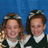 Two people with brown hair stand side-by-side against a blue background, wearing dark jackets over white collared shirts.