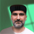 Pixelated static image of a person with short dark hair, light skin tone, and facial stubble wearing a white shirt against green background.
