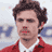 Pixelated static image of a person with dark curly hair wearing a red jacket over a white shirt, set against a blurred background with blue and gray tones.