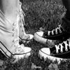 Black-and-white close-up of two pairs of feet wearing sneakers, one pair with white laces and the other with dark laces, positioned on a textured surface.