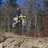 A rider on a white horse gallops across a dirt path in a forested area with blue sky visible.