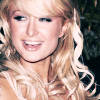 A static image of a person with long, wavy blonde hair, wearing a black choker necklace, and smiling with visible teeth against a dark background.