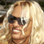 Pixelated close-up of a person with long blonde hair wearing dark sunglasses and a brown top, smiling with visible teeth.