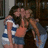 Three people embrace in a close group hug; two wear striped shirts and denim shorts, the third wears a dark top and light shorts.
