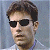 Pixelated close-up of a person with short brown hair wearing dark sunglasses and a light-colored shirt against a blue-gray background.