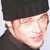 Pixelated close-up of a person wearing a black cap and dark clothing, with light skin and a slight smile, against a light background.
