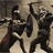 Two stylized figures in dark clothing stand side-by-side against a muted brown background, one holding a long object resembling a sword or staff.