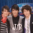 Three young men stand side-by-side against a blue background; the central figure wears a gray jacket with white text and a black shirt underneath.