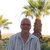 Pixelated image of a person in a light blue shirt standing outdoors with palm trees and a bright sky background.