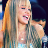 A close-up of a person with long straight blonde hair and bangs, wearing a light blue patterned top and a small pendant necklace, smiling with mouth open.
