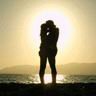 Silhouette of two people embracing at sunset by the water, rendered in low-resolution pixel art with a bright sun behind them.