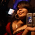 Woman in red dress and blue hat holding a phone, smiling with eyes closed against dark background.