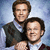 Two men in formal attire pose together against a blue gradient background; one man's arm is visible around the other's shoulder.