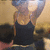 A person in a black tank top with arms raised, standing in front of a blurred background with a bright light source above.