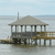 Pixelated static image of a two-story house on stilts over water with a chimney, surrounded by calm water under a light blue sky.