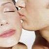 Two people in close proximity with one person kissing the other's cheek, both with closed eyes and visible makeup.