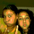 Two people in a yellow top pose closely together, with one person's head slightly turned left and the other facing forward.