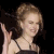 A pixelated image of a person with light brown hair in an updo, wearing a dark spaghetti-strap top and holding a black object near their face against a solid black background.