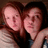 Two people with reddish-brown hair pose closely together, one wearing a light-colored top and the other a dark top, against a dark background.